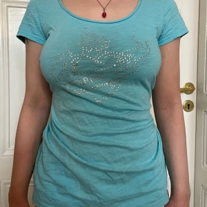 Turkos t-shirt med strassdekor - Snygg turkos t-shirt med rund halsringning och korta ärmar. På första bilden är tröjan knuten i ryggen med en snodd (”orreknut”). Framsidan har ett coolt hjärtmotiv och texten 'ETERNAL LOVE' i glittrande strass.