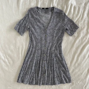 Blommig kortklänning från BikBok - Supersöt blå/vit kortklänning från BikBok med små blommor över hela tyget. Klänningen har v-ringning, knappar framtill och korta ärmar. Längd: 87cm, Midja: 38cm