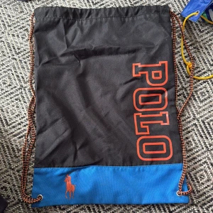 Svart och blå gympåse från Polo Ralph Lauren - Snygg svart och blå gympåse från Polo Ralph Lauren med orange logga och text. Praktisk dragsko och cool design, perfekt för träning eller vardag. Tillverkad i slitstark polyester.