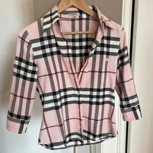 Rutig rosa skjorta från Burberry - Säljer en klassisk rutig skjorta från Burberry i rosa, svart, vit och röd. Skjortan har trekvartsärm, knappar framtill och är tillverkad i 100% bomull. 