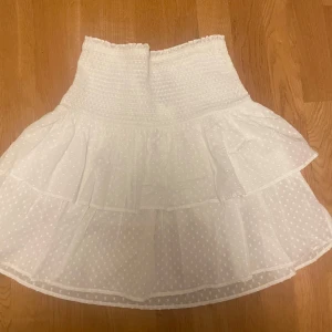 Vit volangkjol med prickigt mönster - Supersöt vit kjol med volanger och diskret prickigt mönster. Kjolen har smock i midjan för en bekväm passform och är tillverkad i ett lätt och luftigt tyg. Perfekt för varma dagar.