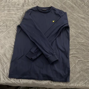 Mörkblå långärmad tröja från Lyle & Scott - Säljer en stilren mörkblå långärmad tröja från Lyle & Scott med klassisk rund hals och den ikoniska gula loggan broderad på bröstet. Perfekt för en clean och enkel look. Materialet känns mjukt och bekvämt.