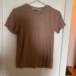 Beige ribbad topp från 157 - Säljer en beige ribbad topp från 157 i storlek S. T-shirten har korta ärmar med en liten volangdetalj och rund halsringning. Perfekt att matcha med jeans eller kjol för en enkel och stilren look.