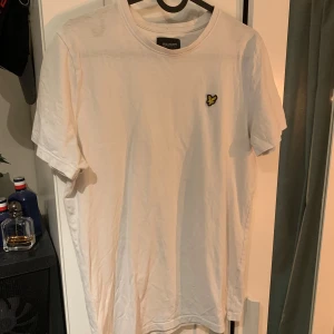 Vit t-shirt från Lyle & Scott  - Säljer en klassisk vit t-shirt från Lyle & Scott  med deras ikoniska gula logga broderad på bröstet. T-shirten har rund hals och korta ärmar. Perfekt för en clean och stilren look. Storlek junior 15/16 år sitter som en S