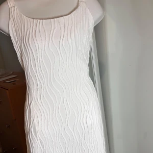 Vit vågigt klänning  - Säljer en elegant vit långklänning med vågigt strukturmönster. Klänningen har breda axelband och rak halsringning, vilket ger en stilren och modern look. Perfekt för sommarens tillfällen eller studentfirande.