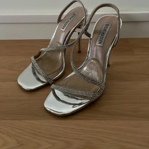 Silverfärgade klackskor från Steve Madden - Säljer ett par glammiga silverfärgade klackskor från Steve Madden med smala remmar dekorerade med glittrande stenar. Skorna har öppen tå och hög smal klack, perfekt för att sticka ut på festen.