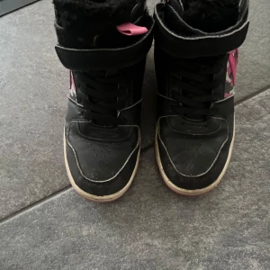 Svarta sneakers med rosa detaljer och fuskpäls - Säljer ett par svarta sneakers med rosa detaljer och fuskpäls på insidan. Skorna har snörning, kardborreband och en platt sula. Perfekta för dig som vill ha både stil och komfort i vardagen.