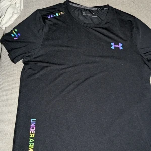 Svart t-shirt från Under Armour Threadborne - Säljer en svart Under Armour Threadborne t-shirt med färgglada loggor och textdetaljer. T-shirten är tillverkad i ett lätt och syntetiskt material, perfekt för träning. Rund halsringning och korta ärmar. Snygg och sportig stil!