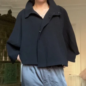 Svart cape-jacka från Zara - Säljer en svart cape-jacka från Zara i storlek L. Jackan har en oversized passform, hög krage och knäppning framtill. Den är tillverkad i ett mjukt material och har en elegant, vid siluett som ger en cool look. Passar mig som är 34