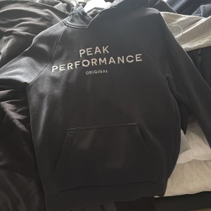 Svart hoodie från Peak Performance - Säljer en svart hoodie från Peak Performance med vit logga på bröstet. Tröjan har huva och en stor magficka framtill. Perfekt för dig som gillar en klassisk och stilren look.