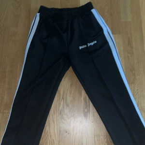 Svarta träningsbyxor från Palm Angels med vita ränder - Palm Angels svarta träningsbyxor med vita sidoremsor och logga på vänster ben. Byxorna har resår i midjan och raka ben, perfekt för en sportig och trendig look.