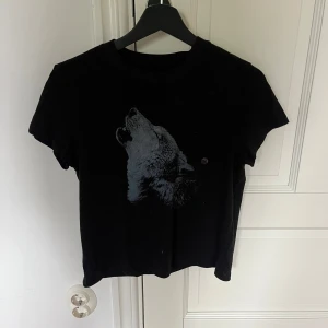 Svart t-shirt med vargtryck från American Eagle - Helt ny med lappar kvar. Svart t-shirt med ett stort tryck av en ylande varg på framsidan. Modellen är croppad och har korta ärmar. Perfekt för dig som gillar djurmotiv och vill ha en cool och avslappnad stil.