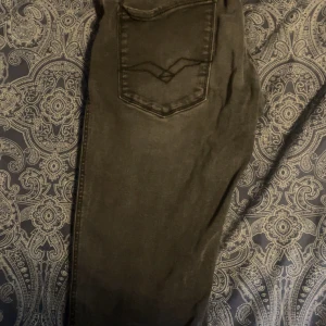 Svarta jeansbyxor från Replay - Säljer ett par svarta jeansbyxor från Replay med klassisk femficksdesign och diskret sliten look. Byxorna har raka ben och normal passform, perfekt till vardags. Snygga detaljer på bakfickan.