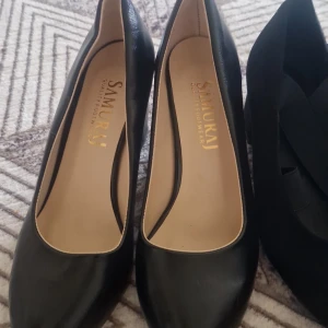 Svarta pumps och ballerinaskor från Samurai - Två par svarta damskor från Samurai. Ena paret är klassiska pumps i slätt skinn med spetsig tå och låg klack. Det andra paret är ballerinaskor i mocka med breda elastiska band över foten och rund tå. Perfekta för en stilren look.
