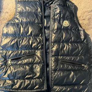 Svart dunväst från Moncler - Snygg svart dunväst från Moncler med glansig finish. Västen har två dragkedjefickor framtill och Moncler-logga på bröstet. Klassisk quiltad design och hög krage. Perfekt för lager-på-lager-stil.ett litet hål på insidan, dm för mer bilder och för frågor🤟🏿🤩👍 