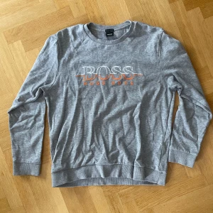 Hugo Boss sweatshirt - Säljer en stilren grå sweatshirt från Hugo Boss med tryckt logga i vitt och orange framtill. Tröjan har rund halsringning och långa ärmar. Storlek M