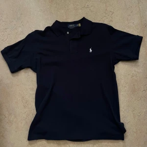 Mörkblå pikétröja från Polo Ralph Lauren - Klassisk mörkblå pikétröja från Polo Ralph Lauren med vit broderad logga på bröstet. Tröjan har krage, knappar vid halsen och korta ärmar. Perfekt för en stilren och avslappnad look.