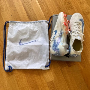 Nike Mercurial fotbollsskor med blå detaljer - Säljer ett par vita Nike Mercurial fotbollsskor med blå glittrig swoosh och orange dobbar. Skorna har snörning och rund tå, och kommer med en vit påse med blå snören.