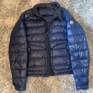 Blå dunjacka från Moncler - Säljer en blå dunjacka från Moncler med klassisk quiltad design och dragkedja framtill. Jackan har lång ärm, två fickor med dragkedja och Moncler-logga på ärmen. Perfekt för kyligare dagar.