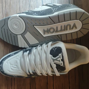 Louis Vuitton sneakers i vitt och grått - Säljer ett par Louis Vuitton sneakers i vitt och grått med klassiskt monogrammönster. Skorna har snörning, rund tå och platt sula. Märkeslogga syns tydligt på sidan och tungan. Perfekta för dig som vill sticka ut med exklusiva sneakers.