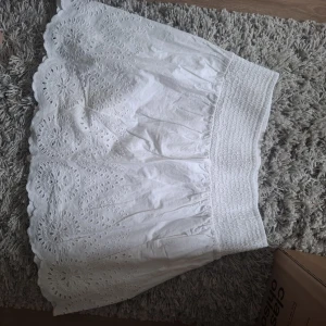 Vit broderad kortkjol med volangkant - Säljer en söt vit kortkjol med bred resår i midjan och broderade blommönster. Kjolen har volangkant nertill och är perfekt för sommarens alla tillfällen. Lätt och luftig känsla.