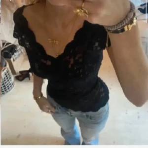 Svart spetsblus med v-ringning - Säljer en svart blus i spets med v-ringning och fina volangdetaljer vid axlarna. Toppen är figurnära och har korta ärmar. Perfekt att styla med jeans eller kjol för en trendig look.