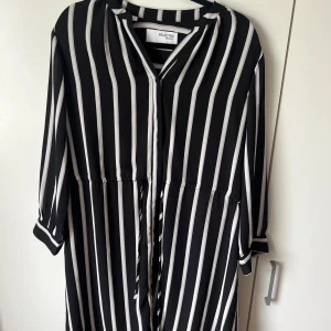 Svartvit randig klänning från Selected Femme - Säljer en svart klänning med vita vertikala ränder från Selected Femme. Klänningen har trekvartsärm, v-ringning och knytband i midjan för en snygg passform. 