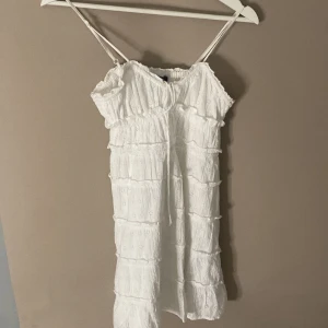 Vit somrig klänning från Gina Tricot - Söt vit klänning från Gina Tricot med smala axelband och volangdetaljer. Klänningen har en luftig och lätt design med broderade detaljer och resår upptill för en bekväm passform. Perfekt för varma dagar.