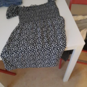 Svart blommig oneshoulder-klänning - Säljer en svart kortklänning med vita små blommor och oneshoulder-design. Klänningen har en smockad överdel och är figurnära upptill med lösare kjol. Perfekt för sommarens alla tillfällen!