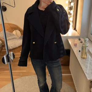 Mörkblå skepparkavaj med guldknappar - Säljer en stilren mörkblå skepparkavaj med bred krage och dubbla rader guldknappar framtill. Jackan har lång ärm och en klassisk, lite kortare passform. Perfekt för dig som vill ha en tidlös look. Ett litet hål i inner tyget i ryggen men absolut inget som syns utifrån 