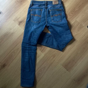 Blå jeans från Nudie Jeans - Säljer ett par klassiska blå jeans från Nudie Jeans med orangea sömmar och snygga bakfickor med vågdetalj. Modellen har normal passform och raka ben. Perfekta för dig som gillar stilrena och tidlösa jeans. Pris kan diskuteras vid snabb affär🤝