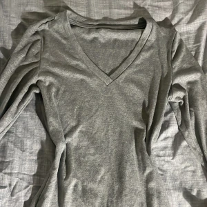 Grå långärmad tröja med v-ringning - Säljer en enkel och stilren grå långärmad tröja med v-ringad hals. Perfekt basplagg som funkar till det mesta och är riktigt skön att ha på sig. Passar dig som gillar en clean och avslappnad stil.