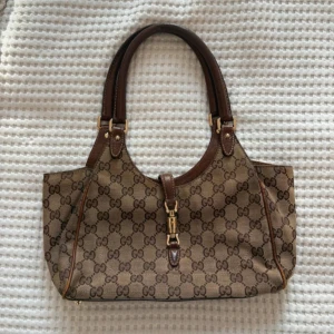 Gucciväska - Säljer en klassisk brun axelväska från Gucci med det ikoniska GG-monogrammet. Väskan har detaljer i brunt skinn, guldfärgade metalldetaljer och ett snyggt spänne framtill. Perfekt storlek för vardagens alla måsten och har dubbla handtag. OBS kan posta fr.o.m 2 juni då jag är på semester tills dess! Har fler bilder så skriv om du vill ha fler.☺️🫶🏼