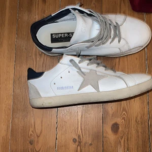 Vita sneakers från Golden Goose med grå stjärna - Säljer ett par vita sneakers från Golden Goose med grå snörning och en grå stjärndetalj på sidan. Skorna har mörkblå häl och vit sula. Perfekta för en avslappnad och trendig stil. Box och påse med följer. Pris kan diskuteras👍