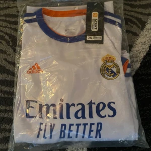 Real Madrid fotbollströja - Säljer en vit Real Madrid fotbollströja från Adidas med blå och orange detaljer. Tröjan kommer med shorts och är helt ny.