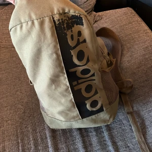 Beige ryggsäck från Adidas - Säljer en beige ryggsäck från Adidas med stort svart logotryck på framsidan. Väskan har meshfickor på sidorna och justerbara axelremmar. Perfekt för skola eller träning och rymmer det mesta du behöver till vardagen.