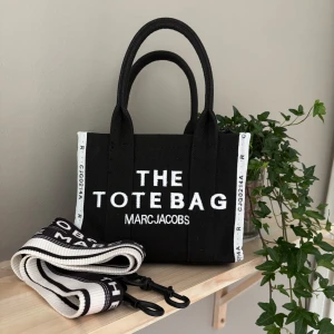 Svart handväska mj the totebag - Säljer en svart handväska  Marc Jacobs ( EJ ÄKTA)  med vit text 'THE TOTE BAG' och 'MARC JACOBS' framtill. Väskan har två handtag och en avtagbar axelrem dock går axelremmen ej att justera. 