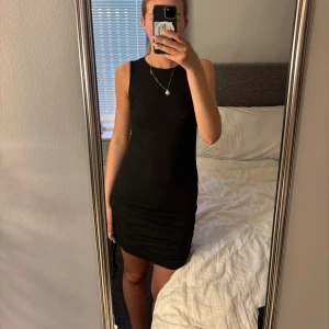 Svart ribbad kortklänning med snörning - Säljer en svart ribbad kortklänning med tight passform och snygg snörningsdetalj på sidan. Klänningen är ärmlös och har en rund halsringning. Perfekt för dig som gillar en enkel men stilren look.