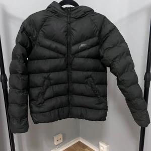 Svart dunjacka från Nike - Säljer en svart dunjacka från Nike med huva och dragkedja framtill. Jackan har två snedställda fickor med knappar och diskret Nike-logga på bröstet. Perfekt för kyliga dagar och riktigt skön att bära.   Jackan har slitage som visas på bilder, den är även sydd på något ställe. Personen som bär jackan är 160 cm lång, passar alltså även en kort XS-S dam.  Jackan säljes i tillfredsställande skick.