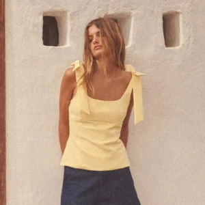 Gul linnetopp med rosettband - Söt ljusgul linnetopp med breda axelband som knyts i stora rosetter på axlarna. Toppen har en fyrkantig urringning och figurnära passform. Perfekt för varma dagar och ger en fräsch sommarkänsla.