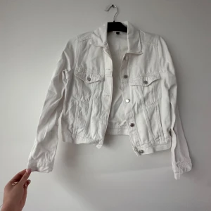 Vit jeansjacka - Säljer en klassisk vit jeansjacka med silverfärgade knappar och två bröstfickor med lock. Jackan har en tidlös design och är perfekt att slänga över en t-shirt eller klänning. Långärmad och i ett mjukt bomullsmaterial.