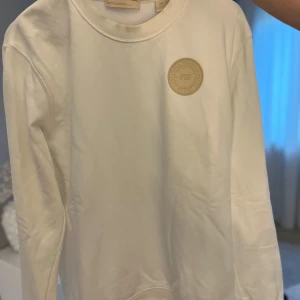 Vit sweatshirt från Scotch & Soda - Säljer en stilren vit sweatshirt från Scotch & Soda med rund halsringning och ett beige broderat märke på bröstet. Tröjan har långa ärmar och en klassisk passform. Perfekt för en clean och avslappnad look. Nypris- 1100kr