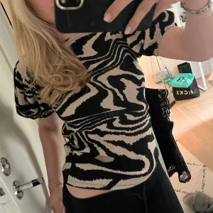 Beige och svart mönstrad topp från Lindex - Säljer en beige och svart topp från Lindex med unikt abstrakt mönster. Toppen har korta puffärmar och är tillverkad i ett stretchigt material som sitter snyggt på kroppen. Perfekt för dig som vill sticka ut lite extra!