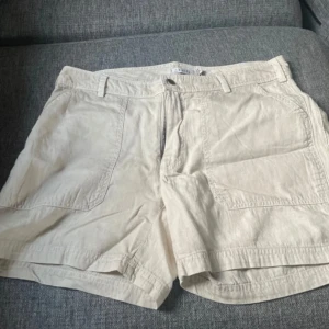 Beiga shorts från Kaffe - Snygga beiga shorts från Kaffe med normal passform. De har stora fickor fram och bak, bälteshällor och stängs med dragkedja och knapp. Perfekta för varma dagar och enkla att matcha med olika toppar.