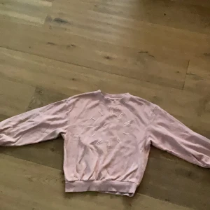 Ljusrosa sweatshirt med broderad text - Säljer en ljusrosa sweatshirt med broderad text 'NEW YORK CITY' i vitt framtill. Tröjan har rund halsringning och långa ärmar. Perfekt för en avslappnad och trendig look.