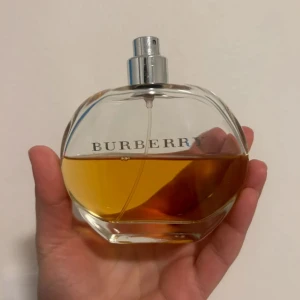 Burberry parfym i rund glasflaska - En elegant parfym från Burberry i en rund, transparent glasflaska med silverfärgad spraykork. Parfymen har en gyllene, bärnstensfärgad vätska och tydlig Burberry-logga på framsidan. Flaskan har en stilren och klassisk design som känns lyxig.