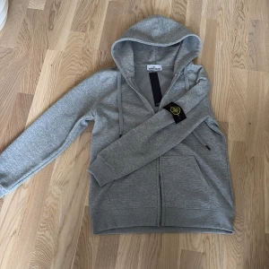 Stone Island kofta - Snygg grå kofta från Stone Island med dragkedja framtill och huva. Tröjan har långa ärmar, ribbade muddar och klassisk Stone Island-logga på ärmen. Perfekt för en avslappnad och stilren look. Priset är inte hugget i sten kan tänka mig sänka vid snabb affär, mer info och bilder privat.