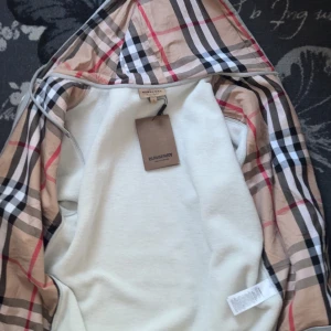 Grå zip-hoodie från Burberry med klassiskt rutigt foder - Säljer en grå hoodie från Burberry med dragkedja och huva. Insidan av huvan har det ikoniska Burberry-rutiga fodret. Broderad logga på bröstet. Perfekt för dig som vill ha en stilren och lyxig look.