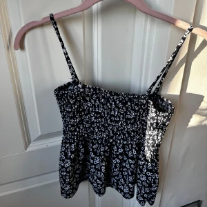 Svart blommig linnetopp från Gina Tricot - Säljer en mörkblå smockad linnetopp med vita små blommor från Gina Tricot. Toppen har tunna axelband och en luftig passform, perfekt för varma dagar. Säljer matchande kjol i en annan ad! Endast använt några gånger ❤️