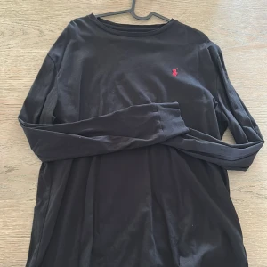 Svart långärmad tröja från Polo Ralph Lauren - Svart långärmad tröja från Polo Ralph Lauren med klassisk röd broderad logga på bröstet. Tröjan har rund halsringning och är tillverkad i mjuk bomull. Perfekt för en stilren och enkel look. Använd fåtal gånger! Inköpt för ca 900kr
